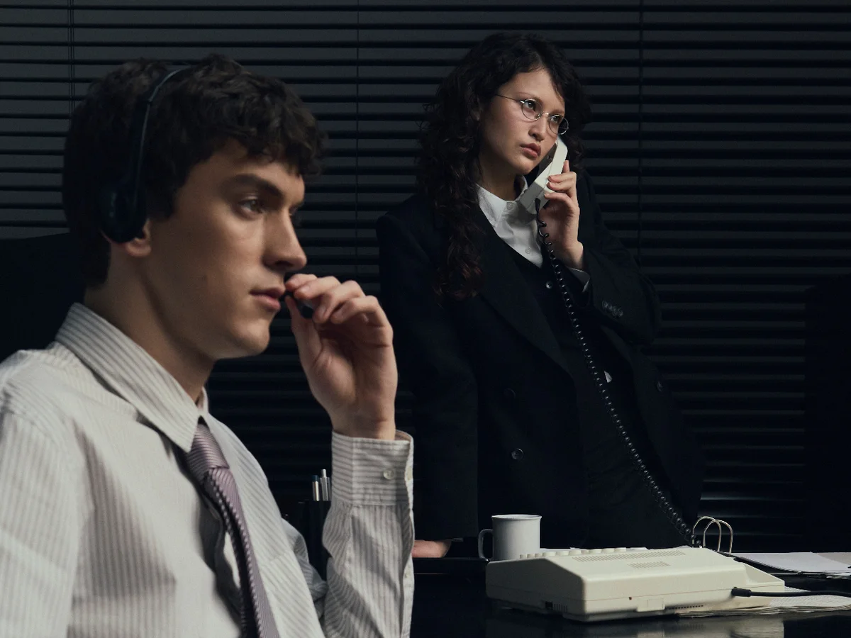 A person wearing a headset beside someone speaking on a corded telephone.